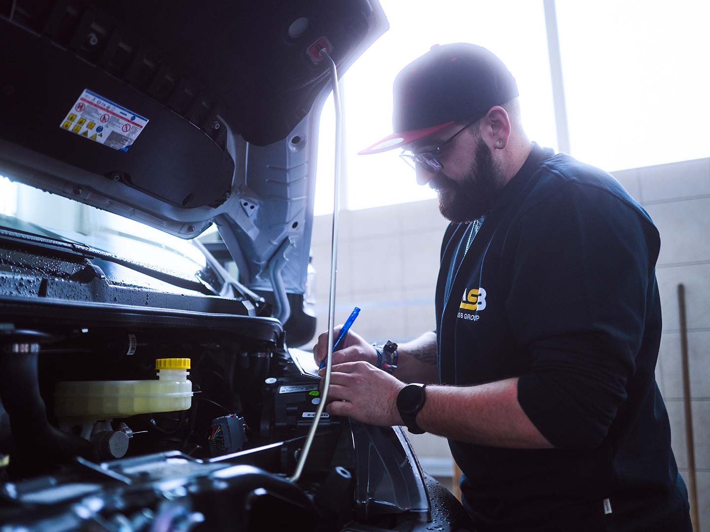 Ein Werkstattmitarbeiter steht an der offenen Motorhaube eines Nutzfahrzeugs und notiert etwas auf einen kleinen Block